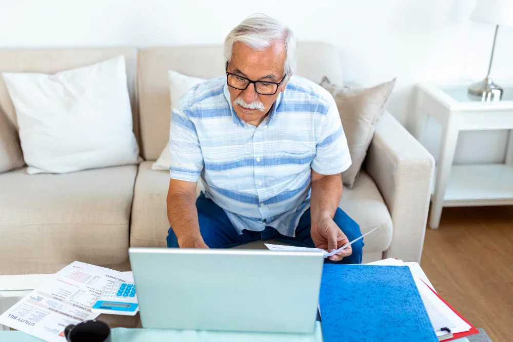 An elderly man sitting on a couch at home, looking concerned while reviewing medical bills and financial documents on a laptop.