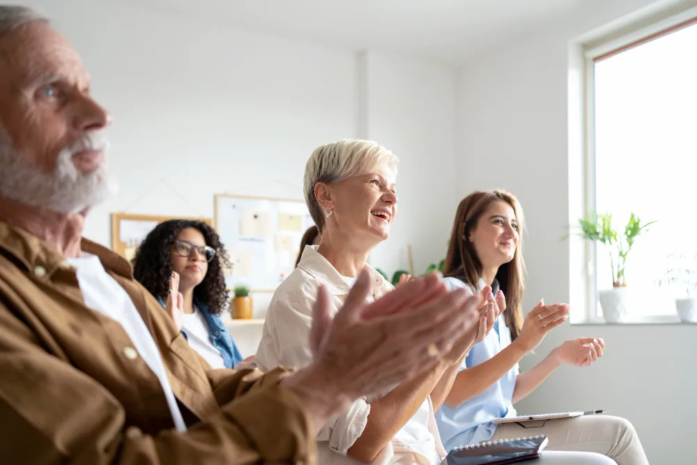 A diverse group of family members and caregivers, including an older man and younger women, clapping and smiling in a supportive community setting.