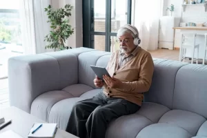 An elderly man with a white beard sits comfortably on a grey sofa in a well-lit ground floor living area, wearing headphones and using a tablet.