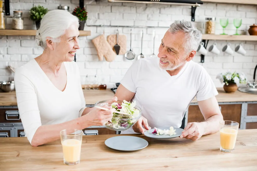 An elderly man and woman, smiling and seated at a kitchen counter with orange juice, share a large, fresh green salad
