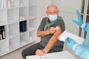 A male patient in a blue shirt sitting in a medical office, while a healthcare professional in blue scrubs and a face mask takes notes on a clipboard during a treatment consultation.