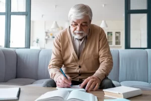 A senior man with a beard and glasses sitting on a sofa, writing in a spiral notebook with open books on the table in front of him.