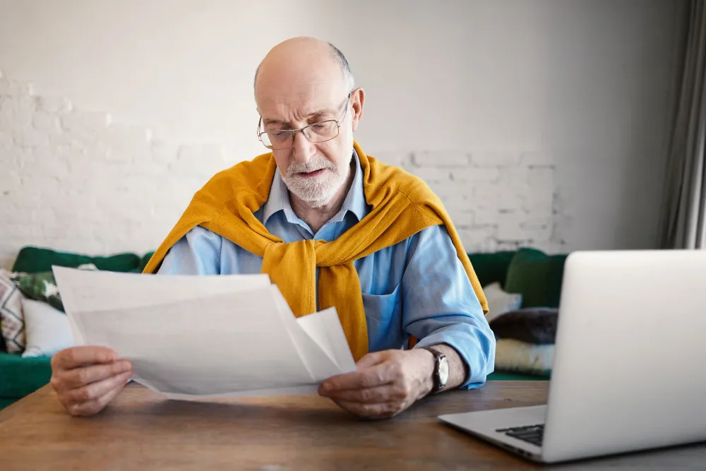 An elderly man wearing glasses carefully reviewing medical and financial documents at a desk with a laptop