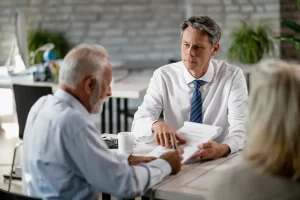 A professional patient advocate in a white shirt and tie explains paperwork to an older couple during a consultation.