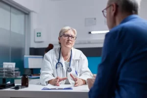 A senior patient in a blue shirt sits across from a female doctor in a white lab coat as she explains medical information with supportive hand gestures.