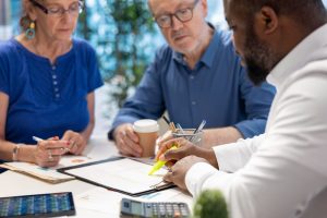 A senior couple sits across a desk from a professional advocate who is pointing to a document with a yellow highlighter during a financial planning meeting.
