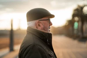 Alt Text: A retired military veteran in a flat cap and winter jacket looking out thoughtfully during sunset, representing a senior seeking earned asbestos benefits.