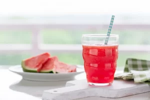 A glass of bright red watermelon juice with a patterned straw sits on a white board next to slices of fresh watermelon.